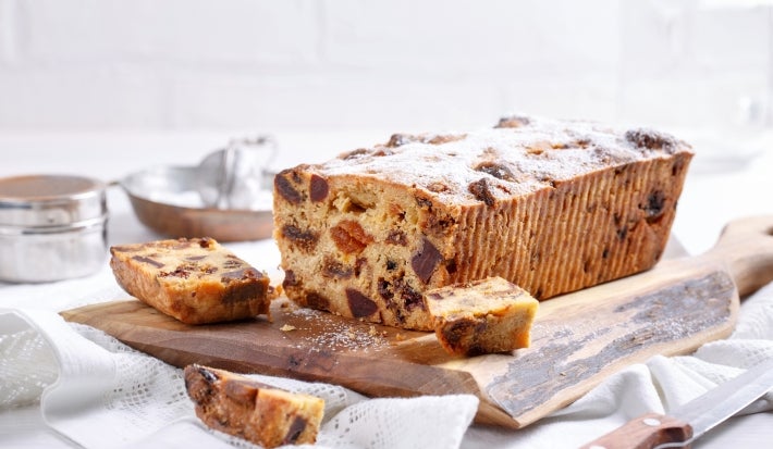 Fotografia de um bolo de frutas recém-assado em uma tábua de madeira, com fatias expostas que mostram sua textura densa e recheio colorido de frutas secas, em uma cozinha iluminada.
