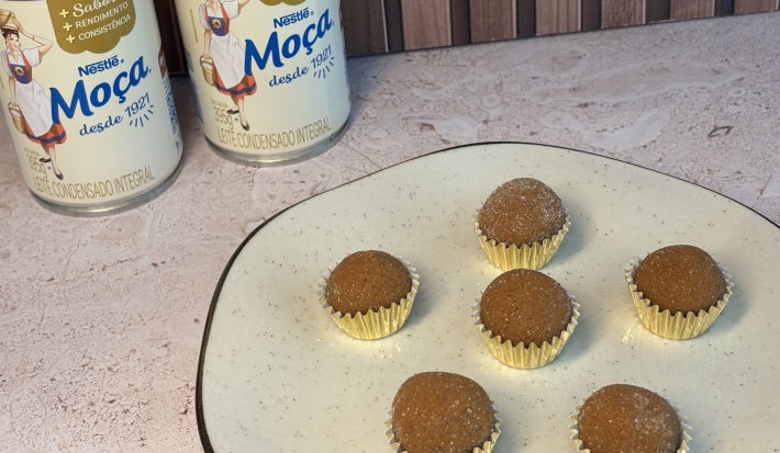 Seis brigadeiros em forminhas douradas sobre prato claro, com duas latas de LEITE MOÇA® ao fundo.