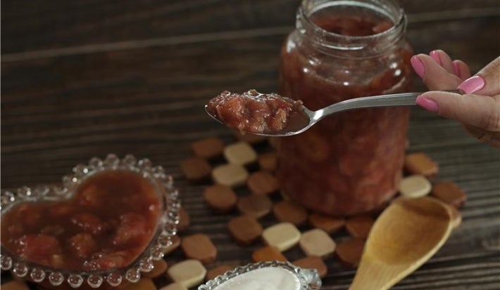 Fotografia em tons escuros de uma mesa de madeira escura, sobre ela tem dois apoios quadriculados de madeira. Apoiado neles tem um pote de vidro com doce de banana, um pote de vidro pequeno em formato de coração e uma mão segurando o doce na colher.