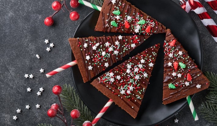 Em cima de uma mesa, prato redondo preto com brownie em forma de triângulo, decorado com ganache e confeitos de bolinhas e estrelas coloridos.