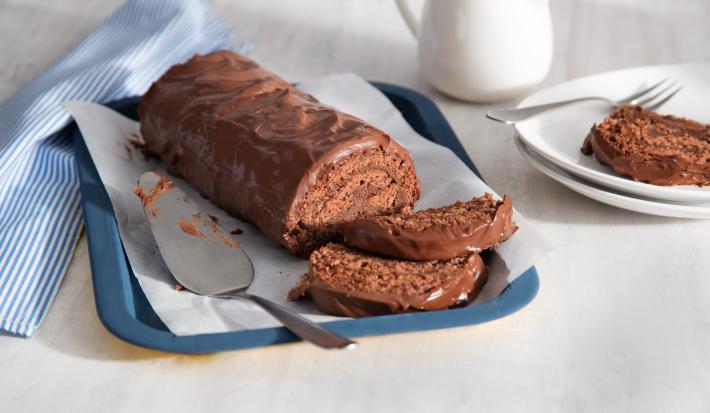 Fotografia em tons de azul, branco e marrom, ao centro da foto o rocambole todo de chocolate com 2 fatias na bandeja azul, ao lado está um prato branco com uma fatia do rocambole e do outro um guardanapo listrado de azul e branco.