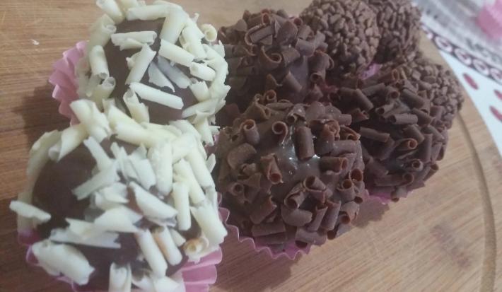Fotografia de oito brigadeiros dentro de forminhas rosa. Seis brigadeiros foram enrolados em confeitos de chocolate preto, e dois deles foram enrolados em confeitos brancos. As forminhas estão sobre uma forma de madeira.