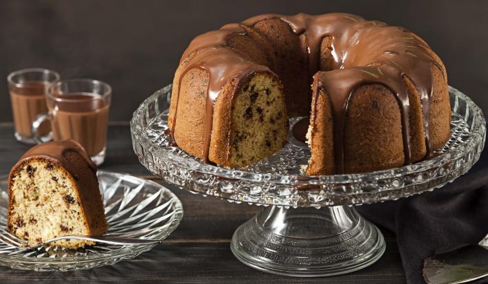 Fotografia em tons de marrom e preto de uma bancada preta vista de frente. Contém um prato redondo transparente com uma fatia de bolo com calda de chocolate por cima, um recipiente transparente para servir o bolo com o bolo sem uma fatia e calda por cima.
