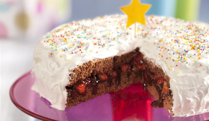 Fotografia em tons de rosa em uma bancada cinza com um suporte para bolo central na cor rosa com o bolo de aniversário confeitado em cima com cobertura branca e confeitos coloridos, cortado ao meio e recheado com chocolate e morango.