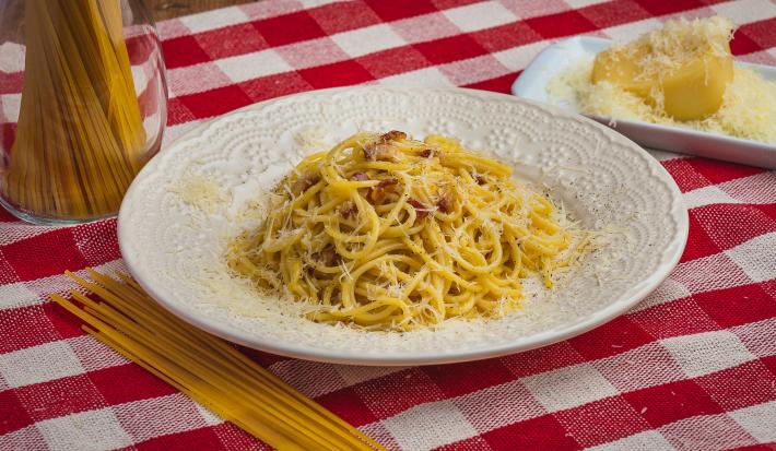 Foto de uma mesa com uma toalha xadrez vermelha e branca. Sobre ela há um prato branco com a receita de Spaghetti à Carbonara, alguns macarrões crus espalhados e num pote e, do lado direito, um pedaço de queijo com queijo ralado. Foto de uma mesa com uma toalha xadrez vermelha e branca. Sobre ela há um prato branco com a receita de Spaghetti à Carbonara, alguns macarrões crus espalhados e num pote e, do lado direito, um pedaço de queijo com queijo ralado.