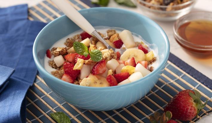 Fotografia em tons de vermelho em uma bancada de madeira de cor branca. Ao centro, um pano azul contendo um bowl azul com as frutas. Ao lado, há um pano azul e ao fundo, um pote com mel e granola.