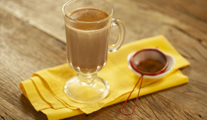 Fotografia em tons de marrom e amarelo de uma bancada de madeira com um paninho amarelo, sobre ele uma xícara transparente com leite, chocolate baton derretido e canela em pó. Ao lado uma peneira vermelha com canela.