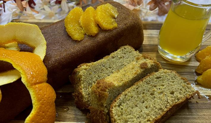 Foto da receita de Bolo de Laranja com Amêndoas. Observa-se um bolo inglês com três pedaços cortados. Como decoração, suco de laranja e a casca cortada.