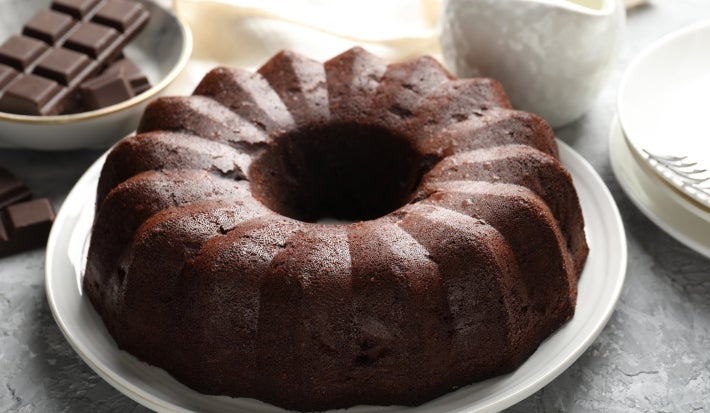 Bolo de chocolate em forma de bundt, com cor marrom escura e superfície brilhante, servido em um prato branco. Ao fundo, um jarro de cerâmica branca e pedaços de chocolate.
