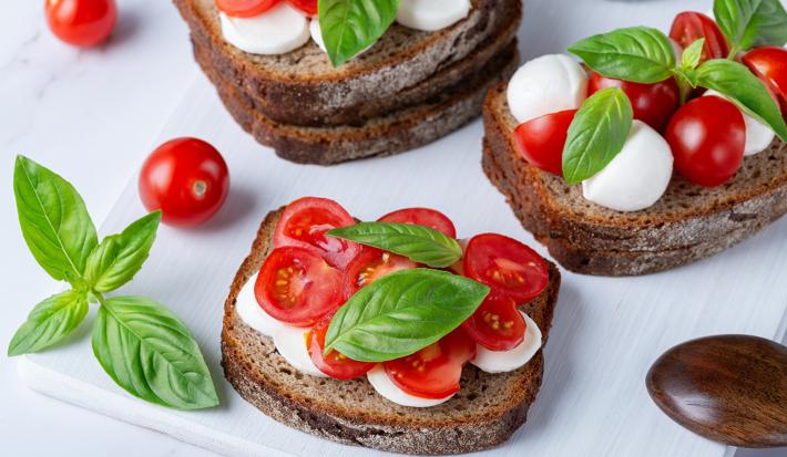 Fotografia de um sanduíche Caprese repleto de cores vibrantes. O pão sustenta as camadas de tomates cereja vermelhos e suculentos, muçarela de búfala macia e folhas de manjericão fresco.