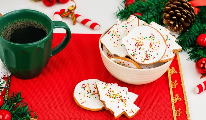 Em cima de uma mesa, tigela redonda com biscoitos em forma de árvore e diversas formas, decorados com glacê branco e confeitos coloridos de bolinha.