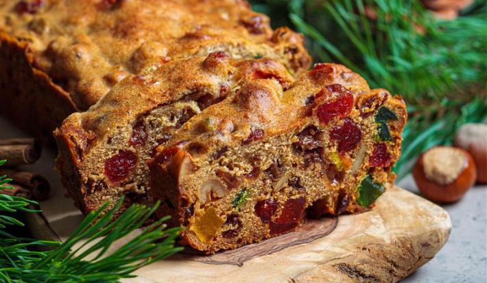 Fotografia de um bolo de frutas cristalizadas e secas com whisky e especiarias, feito na forma pequena de bolo inglês. Tem duas fatias cortadas na frente do restante do bolo, que está sobre um apoio de madeira, ao lado de paus de canela.