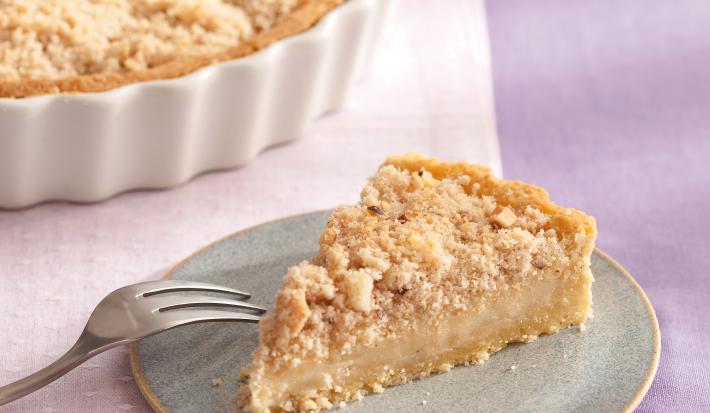 Fotografia em tons de roxo em uma bancada de madeira com uma toalha lilás, um pano rosa, um recipiente branco redondo com a torta. Ao lado, um prato pequeno com a fatia da torta.