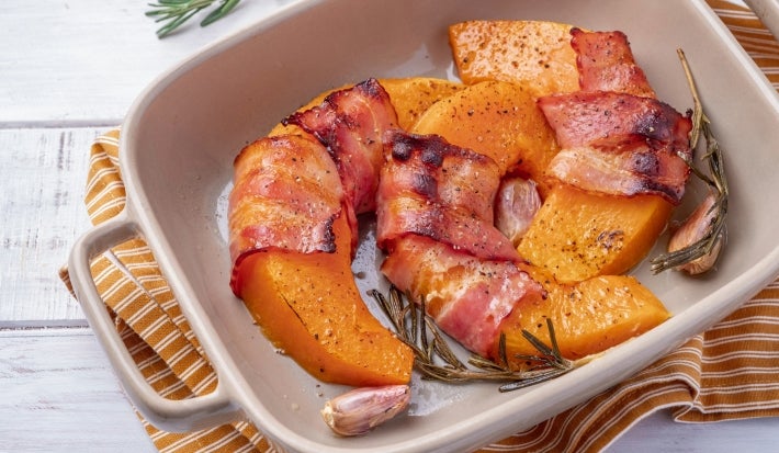 Em cima de uma mesa, há um refratário quadrado com abóboras cortadas em meia-lua enroladas com bacon.
