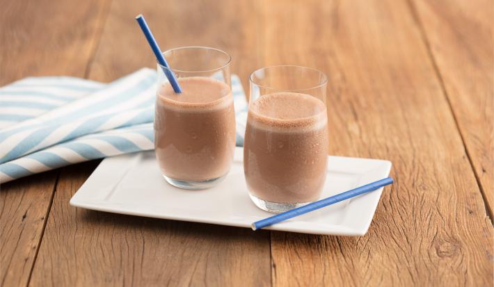 Fotografia em tons de marrom e azul de uma bancada de madeira com um prato branco, sobre ele dois copos de vidro com chocolate gelado e dois canudos azuis. Ao lado um paninho listrado branco e azul.