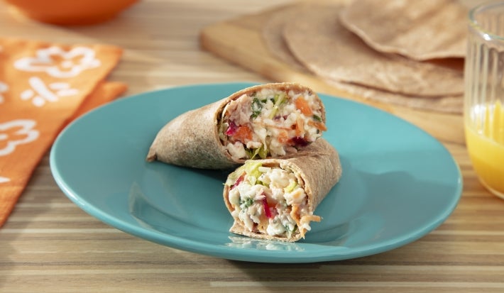 Fotografia mostra wrap cortado ao meio mostrando recheio colorido com beterraba, cenoura e alface, envolto por tortilha integral e pasta cremosa aparente nas bordas. Fotografia mostra wrap cortado ao meio mostrando recheio colorido com beterraba, cenoura e alface, envolto por tortilha integral e pasta cremosa aparente nas bordas.