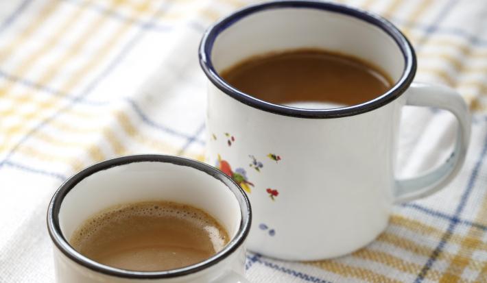 Fotografia em tons de branco, bege e azul, com duas canecas brancas com borda azul contendo bebida em tons café-com-leite, sobre guardanapo branco com listras azul e bege, tudo sobre bancada de madeira.