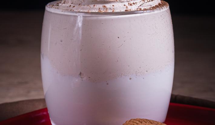 Foto da receita de cappuccino super cremoso servida em um copo de vidro sobre uma louça vermelha em cima de uma mesa de madeira