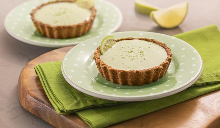 Foto da receita de tortinha de limão. Observa-se dois pratos verdes de sobremesa com as tortinhas sobre um guardanapo verde e uma tábua de madeira. Atrás, duas fatias de limão.