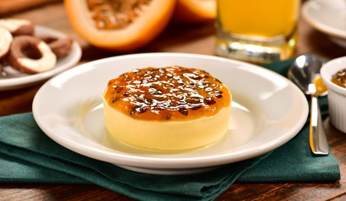 Fotografia de um flan de maracujá com calda por cima, sobre um prato branco e raso branco. Ao fundo, um maracujá cortado na metade, uma jarra com o suco da fruta e alguns recipientes brancos de cerâmica sobre uma mesa de madeira.