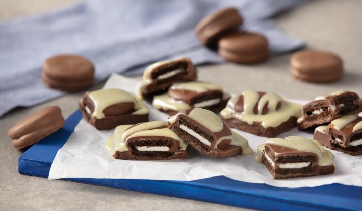 Foto de uma bancada cinza, sobre ela há uma tábua azul com uma folha de papel manteiga. Nela há alguns pedaços cortados da receita pronta além de mais alguns Biscoitos Negresco Coberto espalhados. Ao fundo há um tecido azul claro.