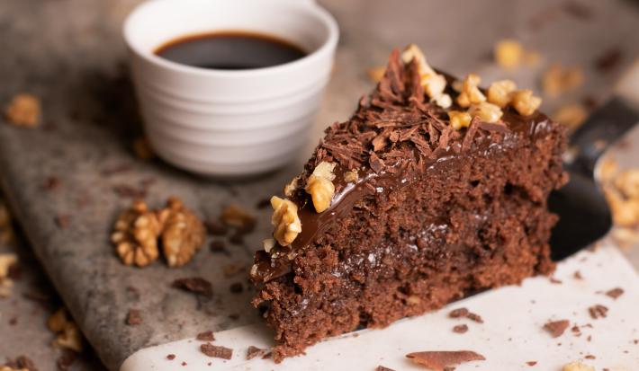 Fotografia de uma fatia de bolo feito com Chocolate NESTLÉ CLASSIC Meio Amargo, nozes e Cobertura de Chocolate ao Leite GAROTO, com raspas de chocolate e castanhas por cima. Ao lado da fatia tem uma xícara pequena branca com café.