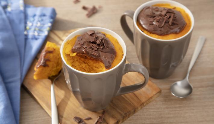 Fotografia em tons de laranja em uma bancada de madeira clara, uma tábua de madeira, um paninho azul, duas canecas marrons com o bolo de cenoura dentro de cada uma e por cima o brigadeiro de Nescau.