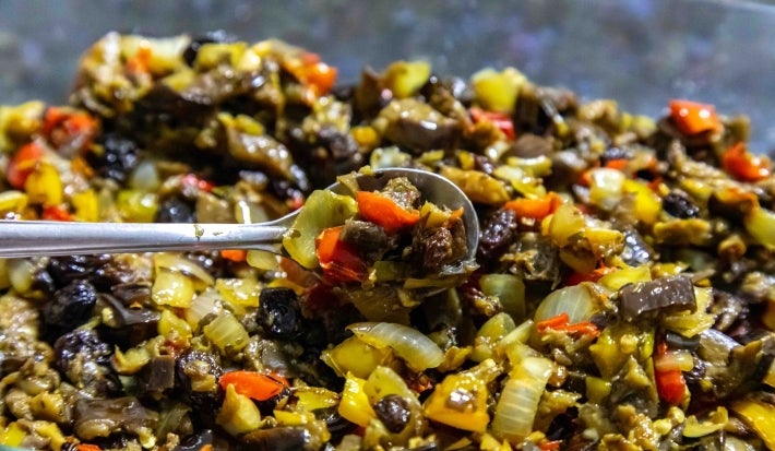 Em cima de uma mesa, colher com berinjelas, pimentões e cebolas picados, regados com azeite.