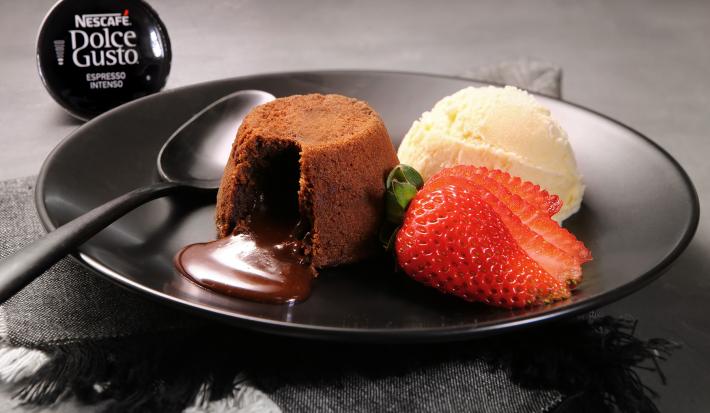 Fotografia em tons de preto em uma bancada de madeira escura, um prato preto redondo com o petit gateau de café em cima, ao lado, uma bola de sorvete de creme e um morango para decorar. Ao fundo, uma cápsula do café intenso de Dolce Gusto.