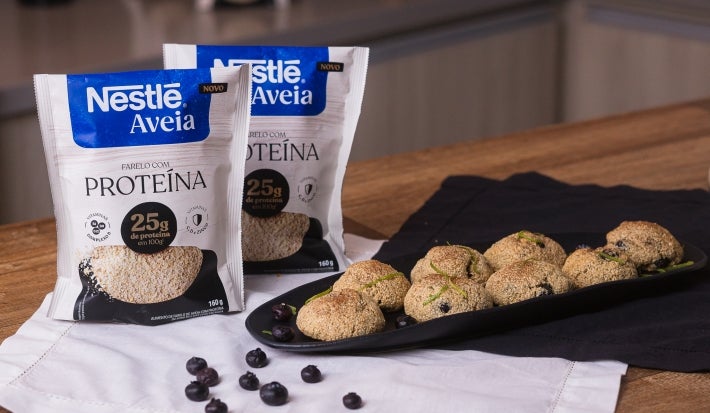 Foto da receita de bolinho de limão e mirtilo com aveia servida em diversas porções sobre uma louça preta em cima de uma mesa de madeira com duas embalagens de nestlé aveia proteína ao lado