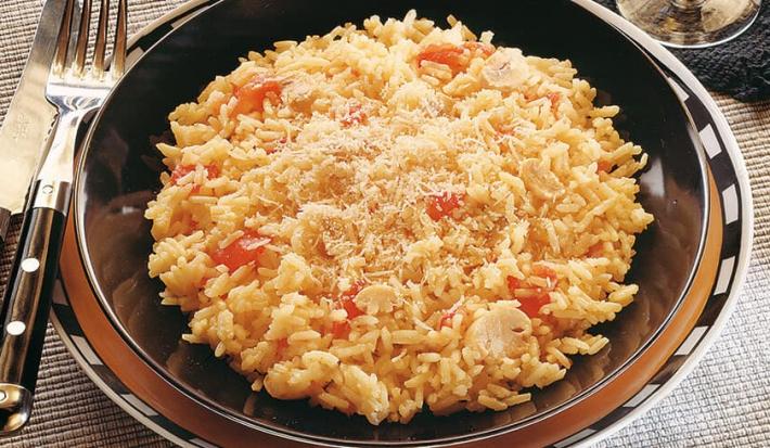 Fotografia em tons de marrom em uma mesa com uma toalha bege, um prato raso marrom, um prato fundo com o arroz à piemontesa dentro dele. Ao lado, talheres e ao fundo, um taça com vinho branco.