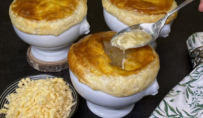 Foto em tons de escuros da receita de sopa de cebola en croute servida em três porções sobre bases de porcelana branca, sendo que há uma colher prateada pegando um pedaço da porção da frente. Ao lado há um prato pequeno com queijo emmenthal ralado