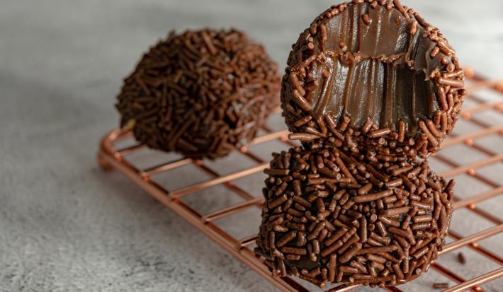 Imagem de três brigadeiros cobertos com chocolate granulado, com um brigadeiro mordido em destaque, mostrando a textura cremosa da receita.
