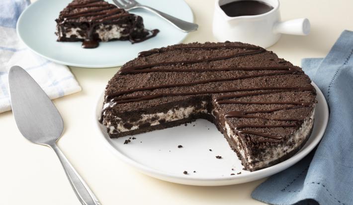 Imagem aproximada da receita de Torta de Sorvete Moça com Calda Quente, servida em um prato branco, sobre uma bancada clara decorada com um tecido azul, um pote branco com calda de chocolate e alguns utensílios