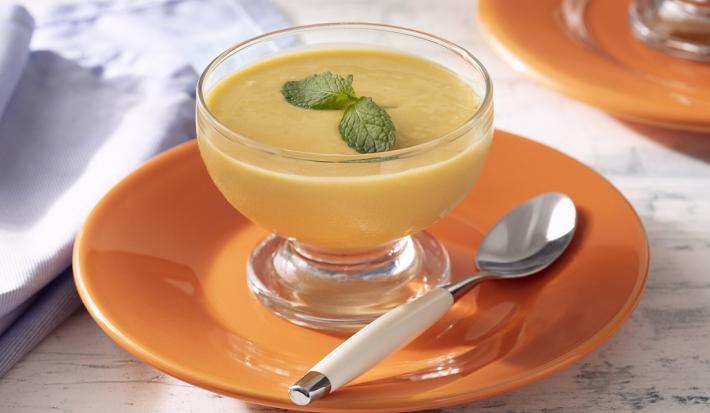 Fotografia em tons de amarelo em uma bancada de madeira de cor branca. Ao centro, um prato laranja com uma taça em cima contendo a mousse e uma colher ao lado. Ao fundo, há um prato laranja e um pano lilás.