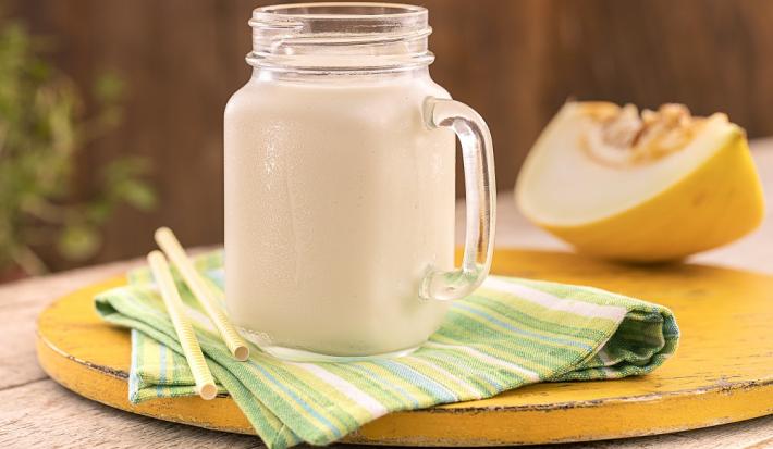 Foto em tons de amarelo da receita de smoothie de melão servida em uma jarra de vidro pequena sobre um pano listrado verde e amarelo com dois canudos ao lado. Ao fundo uma fatia grande de melão