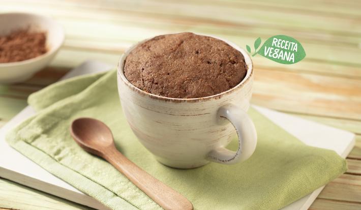 Fotografia em tons de verde, branco e marrom de uma bancada de madeira clara com uma tábua branca e um paninho verde, sobre ele uma caneca branca com o bolo de cacau e uma colher de madeira. Ao fundo um recipiente pequeno redondo branco com cacau em pó.