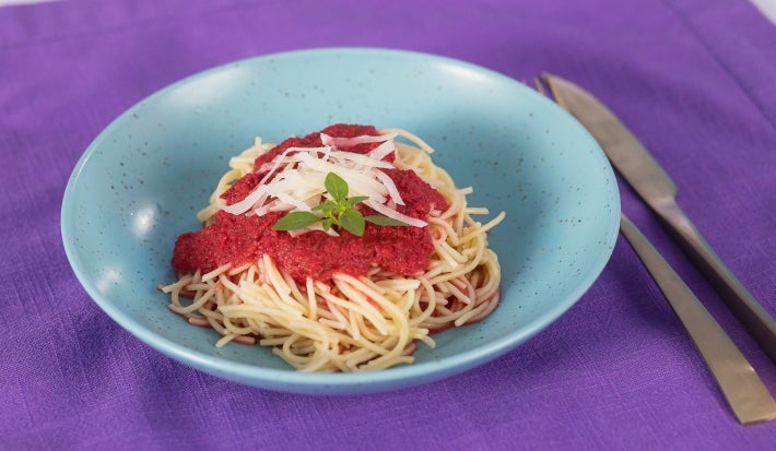 Fotografia em tons claros de um prato de vidro fundo azul sobre um pano roxo. Ao lado tem talheres juntos, como o garfo e a faca, e dentro do prato tem o mcarrão tradicional com o molho de tomate e ascenda.