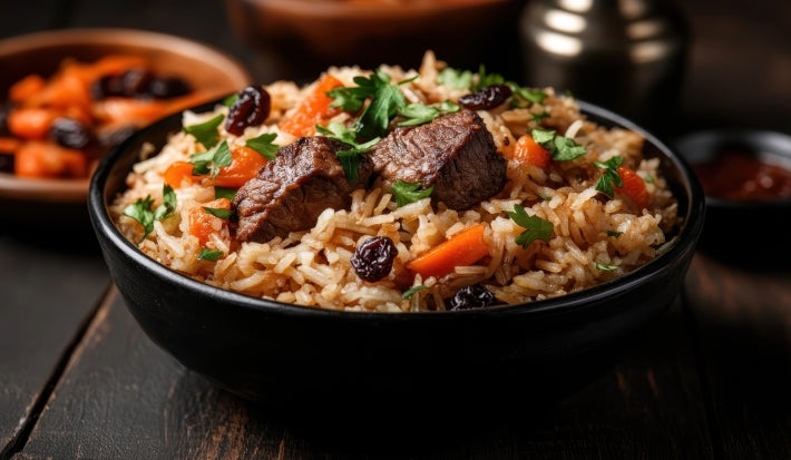 Em um cima de uma mesa, tigela redondo com arroz, carne de boi picada, cenoura e uvas-passas, decorado com cebolinha picada.
