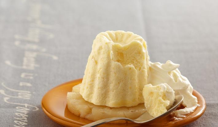 Fotografia em tons de branco em uma bancada de madeira de cor cinza. Ao centro, um prato marrom contendo o flan com uma colher ao lado.