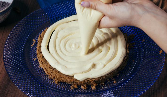 Sobre um prato de vidro de cor azul, há um bolo sendo recheado com um creme de Leite Ninho de cor bege. Para isso, a confeiteira usa um saco de confeitar. Sobre um prato de vidro de cor azul, há um bolo sendo recheado com um creme de Leite Ninho de cor bege. Para isso, a confeiteira usa um saco de confeitar.