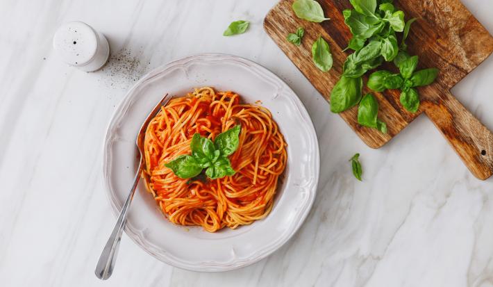 fotografia em tons de branco e vermelho de uma bancada branca vista de cima. Ao centro um prato branco com macarrão espaguete e molho de tomate, e ao lado uma tábua com folhas.