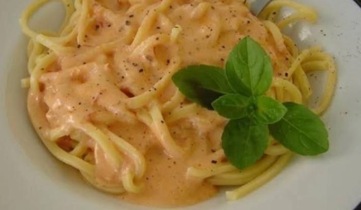 Fotografia em tons de vermelho, em uma bancada de madeira de cor marrom. Ao centro, um prato branco contendo o macarrão.