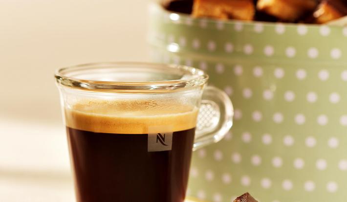 fotografia em tons de marrom, bege e verde tirada de frente de uma xicara transparente com café, e ao fundo um recipiente verde com bolinhas brancas que contém o Caramel Shortcake