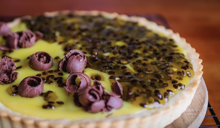 Fotografia da torta de maracujá com base crocante, recheio cremoso e cobertura de geleia natural da fruta. Decorada com raspas de chocolate, traz o contraste ideal entre acidez e dulçor. Uma sobremesa tropical e irresistível!