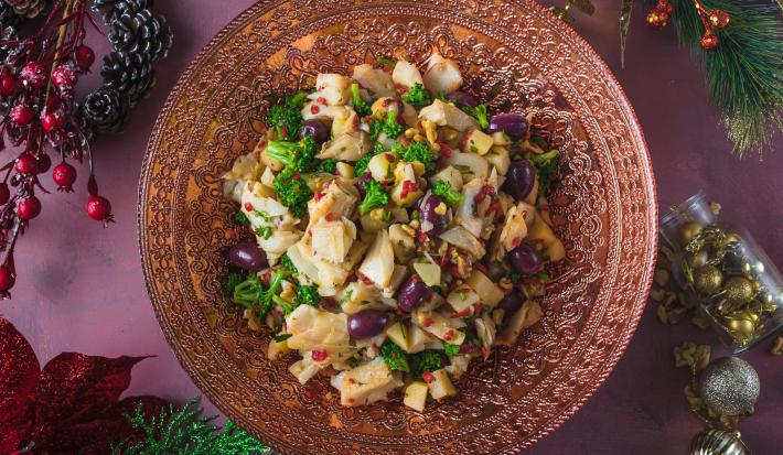 Foto vista de cima da receita de salada de bacalhau em uma travessa redonda vermelha, sobre uma mesa em tom vermelho toda decorada com motivos natalinos ao redor