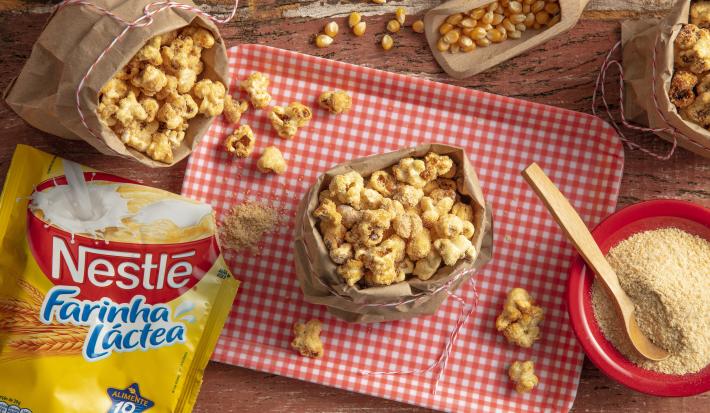 Fotografia em tons de vermelho e amarelo em uma bancada de madeira com uma tábua com listras vermelhas, um saquinho de papel bege com pipocas dentro. Ao lado, uma embalagem de Farinha Láctea Nestlé e um potinho vermelho com farinha láctea.