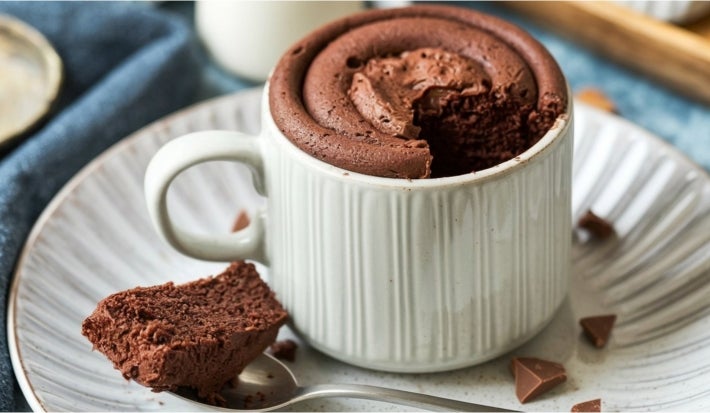Caneca de chocolate em uma caneca branca, com textura fofa e topo levemente rachado. A caneca está sobre um prato branco, com um pedaço do bolo ao lado e uma colher prateada.