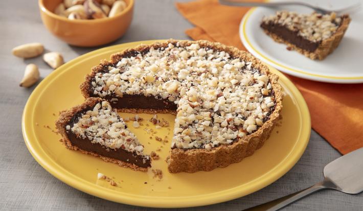 Fotografia em tons de amarelo em uma bancada de madeira de cor cinza. Ao centro, um prato amarelo contendo o cheesecake com uma espátula ao lado. Ao fundo, há um pano laranja com um pires branco em cima contendo uma fatia da sobremesa e algumas castanhas.