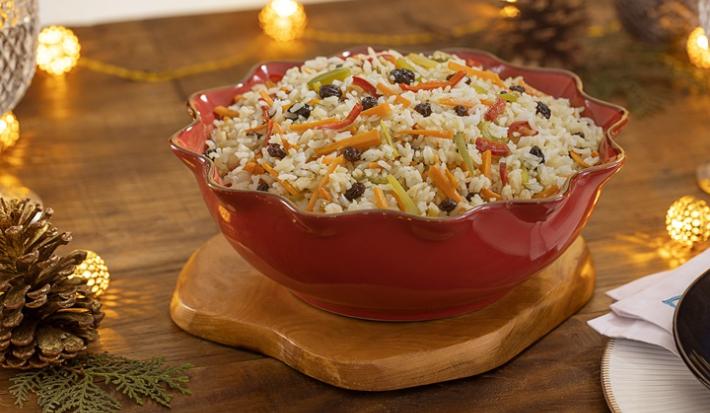 Fotografia mostra um arroz à grega servido em uma travessa. O arroz é soltinho e colorido, com pedaços de cenoura, ervilha, pimentão e uvas-passas distribuídos de forma harmoniosa.